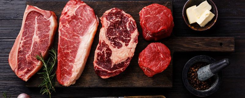 Raw steaks on a dark wooden board beside a carving knife and fork